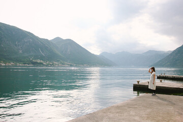 a brunette girl stands alone on the shore and looks into the distance, raising her hand to her face, peering into the sea water or the ocean against the backdrop of a mountain in montenegro