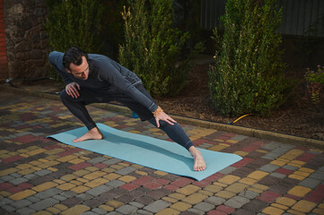 Full length of a man athlete, sportsman standing barefoot on a yoga mat, stretching his legs muscles, practicing a bodyweight training at sunset. Sport. Fitness. Outdoors Workout. Active lifestyle