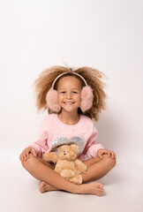 Little child girl wearing earmuffs is hugging teddy bear isolated over white background.