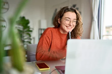Fotobehang Happy young woman using laptop sitting at desk writing notes while watching webinar, studying online, looking at pc screen learning web classes or having virtual call meeting remote working from home. © insta_photos