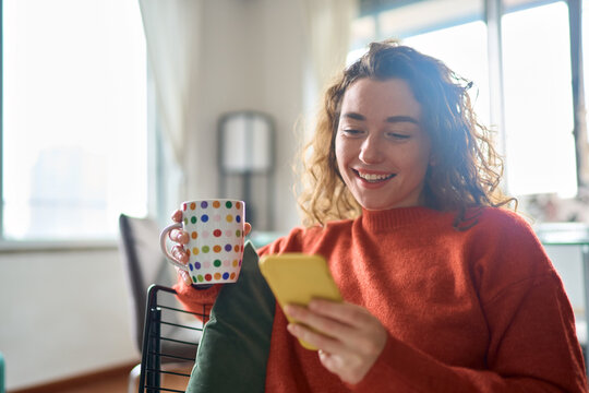 Young happy woman using cell phone drinking coffee relaxing at home. Smiling pretty lady holding smartphone enjoying mobile shopping, checking social media messages, playing game or buying online.