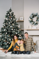 Mom, dad hug daughter and son on floor near Christmas tree. Happy New Year and Merry Christmas. Christmas decorated interior in home. The concept of family holiday. Close up. Winter decor.