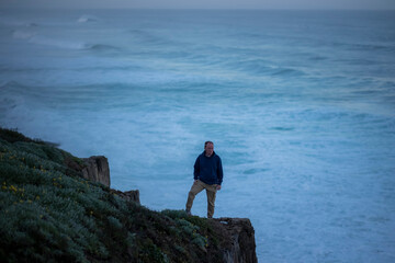 Obraz premium A man stands on the edge of a cliff against the morning ocean surf.