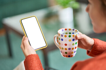 Over shoulder view of woman holding coffee and smartphone with white mock up cellular screen in hands using mobile apps on cell phone. Cellphone display mock up for advertising concept.