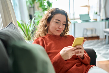 Smiling relaxed young woman sitting on couch using cell phone technology, happy lady holding...