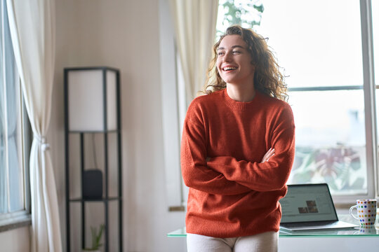 Happy charming smiling confident young adult woman laughing standing in modern cozy apartment interior. Positive pretty lady wearing sweater feeling cheerful looking away at home in living room.