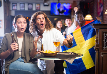 Excited multiracial footbal fans waving the flag of Sweden while drinking beer and watching...