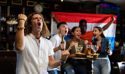 Excited international sport supporters holding up the flag of Netherlands and drinking beer, eating chips in the pub
