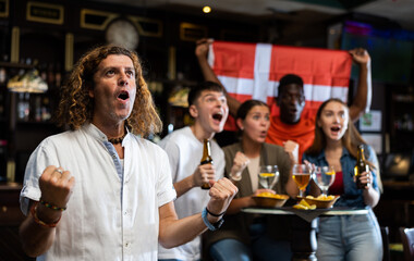 Emotional football fan cheering for team of Denmark while watching match in sports bar with...