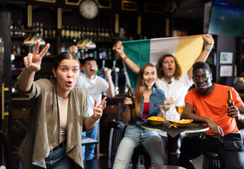 Fototapeta na wymiar Excited young female fan watching sports game in bar, gesturing emotionally, happy with favorite team victory against background of people with Irish national flag
