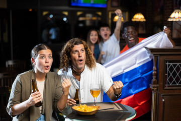 Happy multiracial sport fans holding up in a flag of Russia and watching football competitions in...