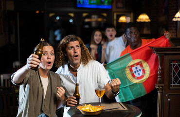 Happy international footbal fans waving the flag of Portugal while drinking beer and watching tournament in sport bar