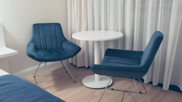 Interior of modern apartment hotel in white and blue colors. Close-up of two comfortable armchairs with round table in cozy hotel room. Vacation at resort. Nobody in the room.