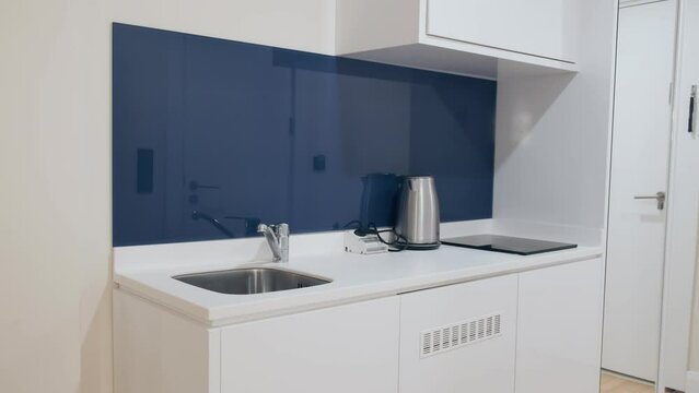 Minimalistic interior of hallway in studio apartment in white and blue shades with white entrance door. Built-in storage and compact kitchen layout. Apartment for one person, student, apart hotel room