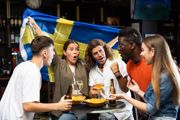 Excited multiracial footbal fans waving the flag of Sweden while drinking beer and watching tournament in the pub