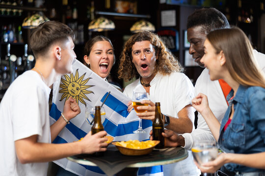 Group of Uruguay football team fans spending time in bar, drinking bear and having fun. People with state flag in pub.