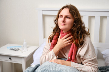 A sick woman holding a sore throat with a red scarf, neck pain. Adult ill woman sitting on a home...
