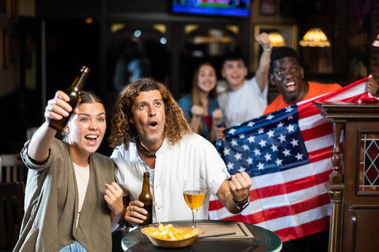 Happy multiracial friends football fans holding the flag of USA, enjoing game, beer and chips in the bar