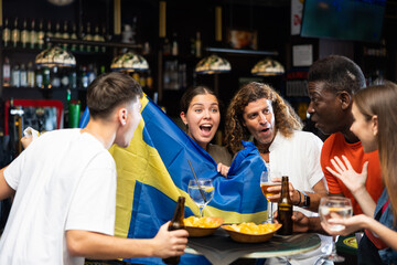 Emotional diverse soccer fans with flag of Sweden, rejoicing winning game with glasses of beer in...
