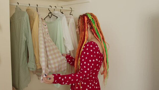 Woman with bright dreadlocks choosing clothes in dressing room. Modern light women's dressing room with various things.