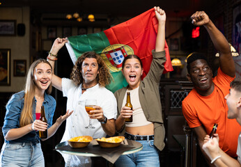 Excited diverse soccer supporters with flag of Portugal watching tournament with pint of beer and...