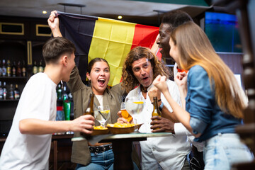 Excited diverse soccer fans with flag of Belgium celebrating victory of team with pint of beer and chips in the pub