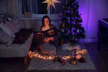 Adult woman reading a book by the Christmas tree in the home living room. Read fairy tales on New Year Eve