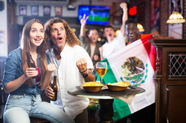 Man and woman, Mexico football team fans, spending time in bar, drinking bear and screaming chants. People with state flag in pub.