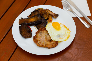 Roasted cutlet with fried egg and frilled vegetables cooked and served on white plate with serving...