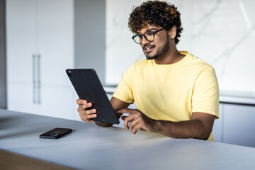 Indian man using digital tablet white sitting in the kitchen