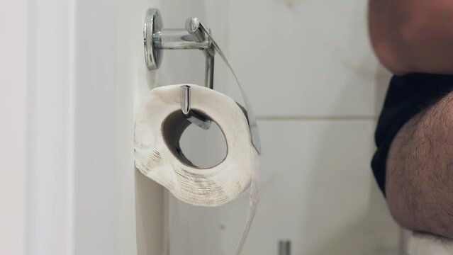 Using toilet paper in a public restroom. Wall mounted metal bumf holder. Man sitting on toilet next to roll dispenser. Unwinding and tearing off sanitary paper.