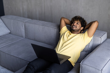 Satisfied relaxed young indian man chilling on sofa in living room, smiling. Handsome guy have rest at home, after laptop distant work, dreaming about vacations, throwing his hands behind