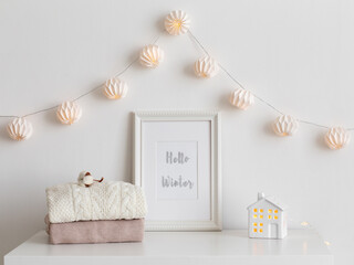 Frame with text HELLO WINTER, knitted sweaters, decorative ceramic house and paper garland on white table. Christmas cozy winter home decor. New year interior decorations.