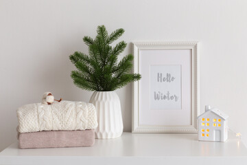 Frame with text HELLO WINTER, fir branch in vase, knitted sweaters and decorative ceramic house on white table. Christmas cozy winter home decor. New year interior decorations.