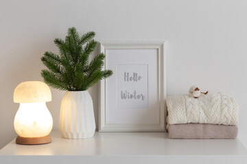 Frame with text HELLO WINTER, fir branch in vase, knitted sweaters and night lamp on white table. Christmas cozy winter home decor. New year interior decorations.