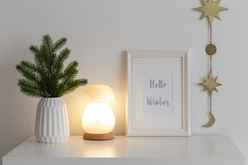 Frame with text HELLO WINTER, fir branch in vase, night lamp and golden garland with stars on white table. Christmas cozy winter home decor. New year interior decorations.