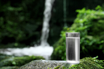 Glass of clean water near mountain waterfall outdoors. Space for text