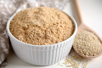 Sesame flour in bowl and wooden spoon of seeds on white table, closeup