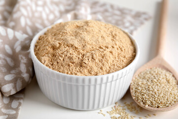 Sesame flour in bowl and wooden spoon of seeds on white table, closeup