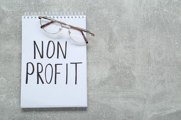 Notebook with phrase Non Profit and glasses on light grey table, flat lay. Space for text