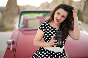 Fototapeta na wymiar beautiful girl in retro style posing near vintage red cabriolet car with old film camera in hands.