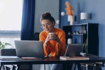 Beautiful woman holding coffee cup and using laptop while sitting at her working place in office