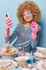 Vertical shot of curly haired housewife washes dirty dishes after party poses at smartphone front camera wears rubber gloves applies hydrogel patches under eyes wears festive dress blue background