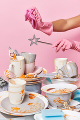 Cropped shot of unknown person in rubber protective gloves holds magic wand and cloth over pile of untidy dishes on table against pink background. Household and home hygiene routine concept.