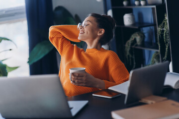 Happy young woman holding coffee cup and keeping her feet on desk while sitting in office