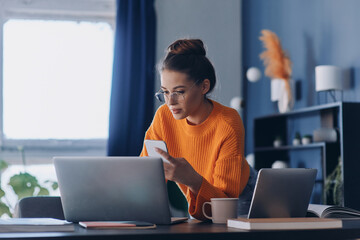 Beautiful young woman holding mobile phone and looking at laptop while working in office