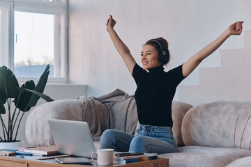 Excited young woman in headphones keeping arms raised while sitting near the laptop at home