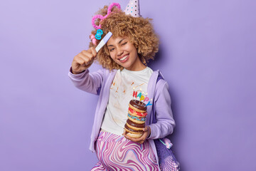 Motherhood pregnancy concept. Positive pregnant woman with curly hair holds positive test and delicious appetizing doughnuts celebrates birthday dressed casually isolated over purple background