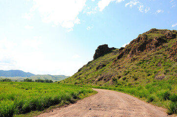 A dirt field road turns at the foot of a high hill with rocky formations at the top.