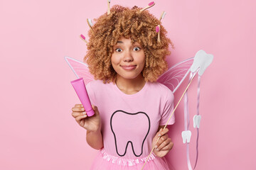 Horizontal shot of curly haired tooth fairy holds tube of toothpaste and wand smiles pleasantly wears pink t shirt tells how to care about teeth isolated over rosy background. Oral hygiene concept